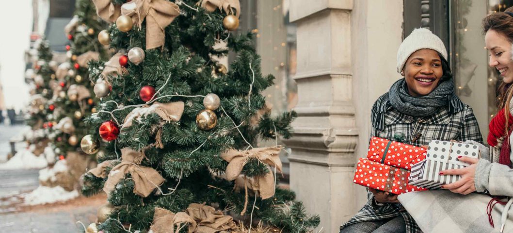 two women exchanging gifts in winter clothes on a city sidewalk.